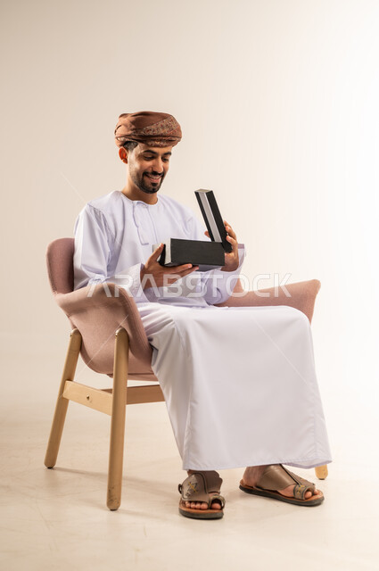 مفهوم الشكر والامتنان، تقديم الهدايا بالاعياد والمناسبات الخاصة، التعبير عن الحب والتقدير والاحترام، بورتريه لرجل عربي خليجي عماني يرتدي الدشداشة والعمامة يجلس على أريكة مريحة يقوم بفتح صندوق الهدية بايماءات السرور والسعادة، خلفية بيضاء
