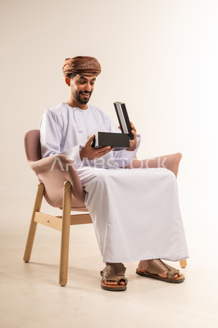 تقديم الهدايا بالاعياد والمناسبات الخاصة، مفهوم الشكر والامتنان، التعبير عن الحب والتقدير والاحترام، بورتريه لرجل عربي خليجي عماني يرتدي الدشداشة والعمامة يجلس على أريكة مريحة يقوم بفتح صندوق الهدية بايماءات المفاجئة والصدمة، خلفية بيضاء