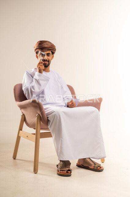 Concept of examination and exploration, image clarification and close-up view, portrait of an Arab Gulf Omani man wearing a dishdasha and turban sitting on a comfortable chair holding a magnifying glass in his hand, placing it on his eye with gestures of concentration and contemplation, white background
