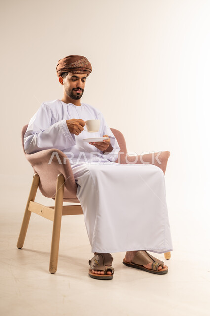 تناول الأعشاب الشتوية الدافئة، الاستمتاع بالمشروبات الساخنة، قضاء وقت الفراغ باحتساء المشروب المفضل, بورتريه لرجل عربي خليجي عماني مبتسم يرتدي الدشداشة والعمامة يجلس على اريكة مريحة يقوم بشرب القهوة بإيماءات السعادة والسرور، خلفية بيضاء