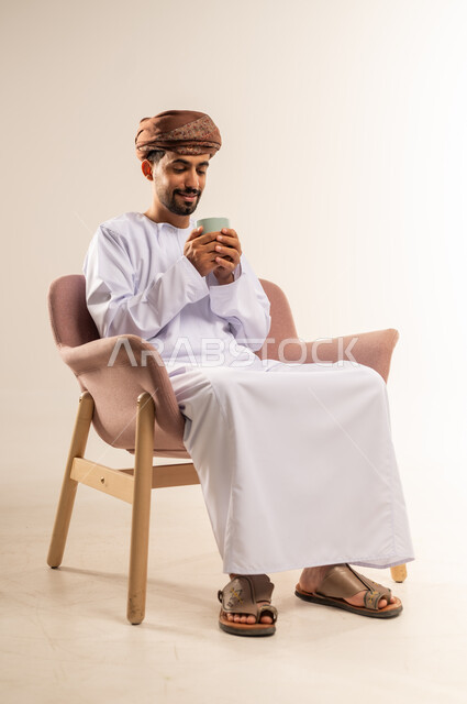 قضاء وقت الفراغ باحتساء المشروب المفضل, تناول الأعشاب الشتوية الدافئة، الاستمتاع بالمشروبات الساخنة، بورتريه لرجل عربي خليجي عماني مبتسم يرتدي الدشداشة والعمامة يجلس على اريكة مريحة يقوم بشرب القهوة بإيماءات السعادة والسرور، خلفية بيضاء