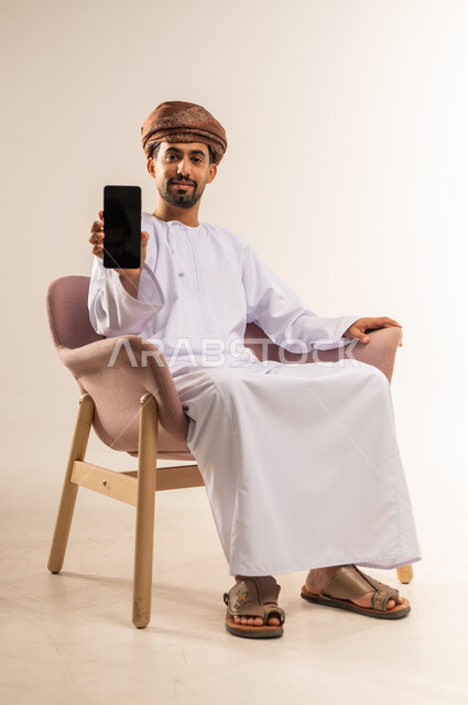 عرض شاشة  سوداء فارغة، استخدام التقنيات الحديثة والأجهزة المتطورة، بورتريه لرجل عربي خليجي عماني يرتدي الدشداشة والعمامة يمسك بيده الهاتف المحمول، النظر إلى الكاميرا بإيماءات السعادة والسرور، إعلان ودعاية ترويجية، خلفية بيضاء