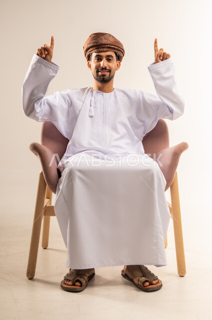 حركات يدين تدل على هذا المكان (هنا)، تحديد موقع شيء ما، بورتريه لرجل عربي خليجي عماني مبتسم يرتدي الدشداشة والعمامة يجلس على أريكة مريحة يشير بأصبعي السبابة نحو الاعلى بايماءات السرور والسعادة، خلفية بيضاء