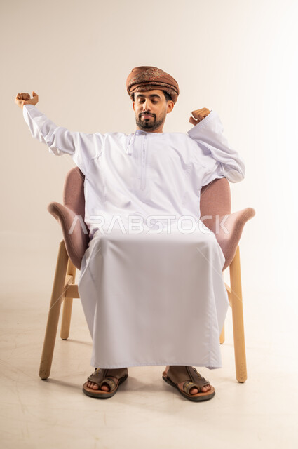الاحساس بالتعب والارهاق، بورتريه لرجل عربي خليجي عماني يرتدي الدشداشة والعمامة يجلس على كرسي مريح يرفع يديه الى الاعلى بإيماءات النعاس، الشعور بالكسل والخمول، الحاجه الى النوم والراحه، خلفية بيضاء