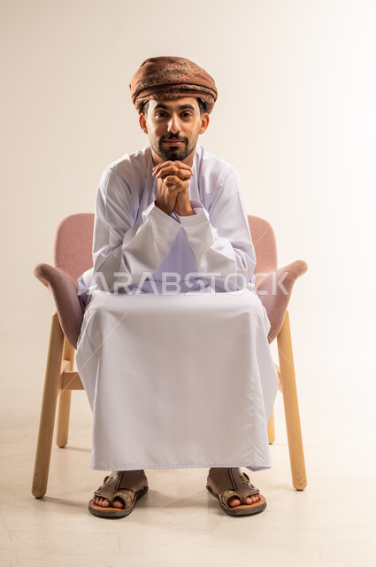 تعابير الثقة بالنفس، مفهوم الرجولة والوقار، الاهتمام بالمظهر الخارجي، بورتريه لرجل عربي خليجي عماني يرتدي الدشداشة والعمامة يجلس على كرسي مريح يضع يديه أسفل ذقنه بايماءات السرور، خلفية بيضاء