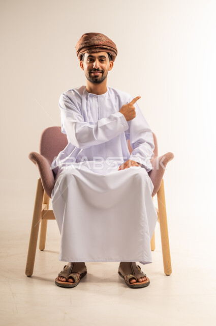 تحديد موقع شيء ما، اشارات يدين تدل على هذا المكان (هنا)، بورتريه لرجل عربي خليجي عماني مبتسم يرتدي الدشداشة والعمامة يجلس على أريكة مريحة يشير بأصبعي السبابة نحو اليسار بايماءات السرور والسعادة، خلفية بيضاء