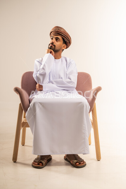 Finding a solution to a problem, contemplation and planning for the future, portrait of an Arab Gulf Omani man wearing a dishdasha and turban, putting his hand on his chin, looking somewhere with gestures of thinking and concentration, expressions of absent-mindedness and contemplation, white background