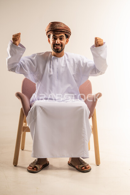 القوة والتحدي والحماس، مفهوم الانجاز وتحقيق الاهداف، بورتريه لرجل عربي خليجي عماني يرتدي الدشداشة والعمامة يجلس على كرسي مريح يرفع قبضتيه نحو الأعلى بإيماءات التشجيع، تعابير السرور والسعادة، خلفية بيضاء