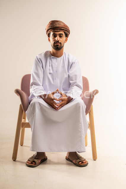 مفهوم الرجولة والوقار، تعابير الثقة بالنفس، الاهتمام بالمظهر الخارجي، بورتريه لرجل عربي خليجي عماني يرتدي الدشداشة والعمامة يجلس على كرسي مريح بايدي متشابكة ينظر الى الكاميرا بايماءات الجدية، خلفية بيضاء