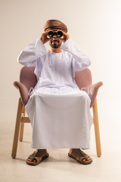 رؤية الاشياء البعيدة وتقريبها، حب الاستكشاف والمغامرة، بورتريه لرجل عربي خليجي عماني يرتدي الدشداشة والعمامةيجلس على كرسي مريح يحمل بيده منظار ثنائي العينين ينظر بإيماءات السرور والاستمتاع، خلفية بيضاء
