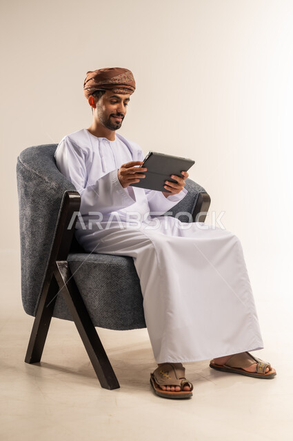 الدردشة مع الأهل والأصدقاء، استعمال التكنولوجيا والتقنيات الحديثة، بورتريه لرجل عربي خليجي عماني يرتدي الدشداشة والعمامة يحمل بيده جهاز التابلت، تصفح مواقع التواصل الاجتماعي، استخدام تطبيقات وبرامج الأجهزة اللوحية، خلفية بيضاء