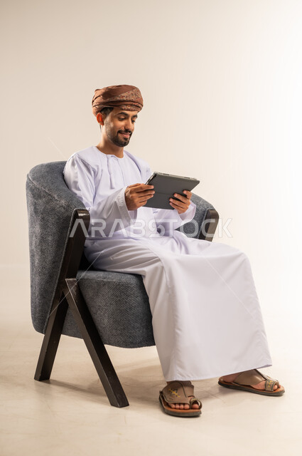 استعمال التكنولوجيا والتقنيات الحديثة، الدردشة مع الأهل والأصدقاء، بورتريه لرجل عربي خليجي عماني يرتدي الدشداشة والعمامة يحمل بيده جهاز التابلت، تصفح مواقع التواصل الاجتماعي، استخدام تطبيقات وبرامج الأجهزة اللوحية، خلفية بيضاء