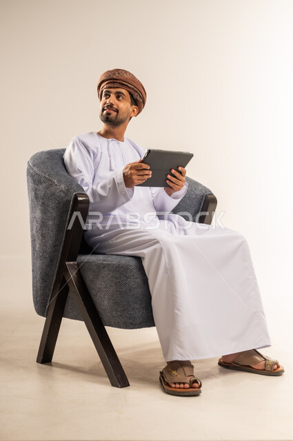 Browsing social media, using technology and modern techniques, chatting with family and friends, portrait of an Arab Gulf Omani man wearing a dishdasha and turban holding a tablet in his hand, looking at a place with focus gestures, using tablet applications and programs, white background