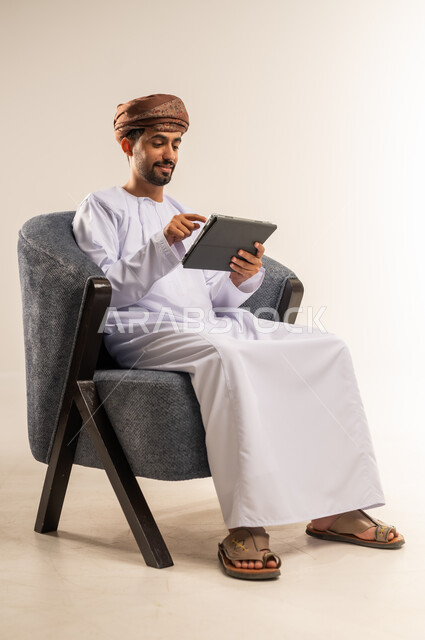 الدردشة مع الأهل والأصدقاء، استعمال التكنولوجيا والتقنيات الحديثة، بورتريه لرجل عربي خليجي عماني يرتدي الدشداشة والعمامة يحمل بيده جهاز التابلت، تصفح مواقع التواصل الاجتماعي، استخدام تطبيقات وبرامج الأجهزة اللوحية، خلفية بيضاء