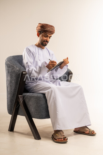 اعداد تقارير ومهام العمل، مراجعة نتائج الاجتماع، بورتريه لرجل عربي خليجي عماني يرتدي الدشداشة والعمامة يجلس على كرسي مريح يحمل بيده كراسه تدوين الملاحظات يقوم بتصفح الاوراق، دراسة جدوى الاعمال، خلفيه بيضاء