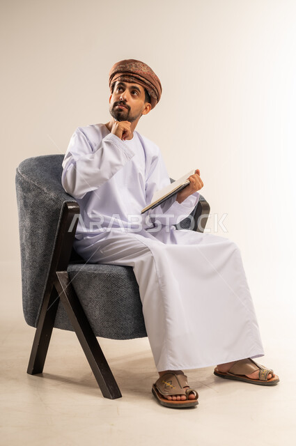 دراسة جدوى الاعمال، اعداد تقارير ومهام العمل، مراجعة نتائج الاجتماع، بورتريه لرجل عربي خليجي عماني يرتدي الدشداشة والعمامة يجلس على كرسي مريح يحمل بيده كراسه تدوين الملاحظات يضع يده اسفل ذقنه بايماءات السرور يقوم بتصفح الاوراق، خلفيه بيضاء
