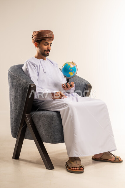 مفهوم حب البحث واستكشاف القارات في الجغرافيا, بورتريه لرجل عربي عماني خليجي يرتدي الدشداشة والعمامة يحمل بيده مجسم الكرة الارضية, تعابير السرور و الفرح بالانتماء للدول العربيه, خلفيه بيضاء
