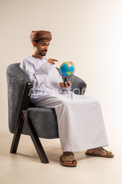 Pointing to the globe, portrait of an Omani Gulf Arab man wearing a dishdasha and turban pointing with his index finger to the world map, expressions of pleasure and joy at belonging to the Arab countries, the concept of love of research and exploration of continents in geography, white background