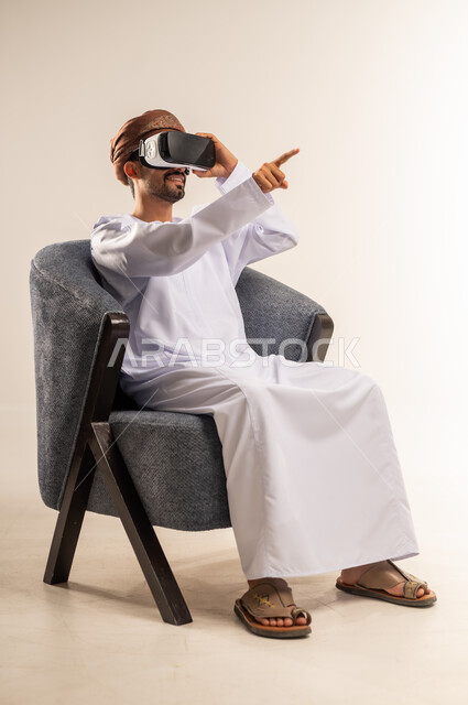 Integration and simulation of the imaginary world, fun and entertainment by watching through virtual reality glasses, portrait of a young Arab Gulf Omani man wearing a dishdasha and turban sitting on a comfortable chair looking through 3D glasses pointing to a place with gestures of pleasure, watching realistic movies, white background