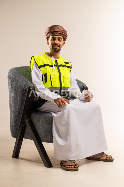 Working in the engineering sector, the concept of engineering and architectural construction, a portrait of an Arab Gulf Omani engineer wearing a dishdasha, turban and a protective vest sitting on a comfortable chair looking at the camera with gestures of pleasure and happiness, white background