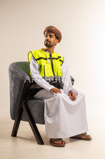 مفهوم الهندسة والانشاءات المعمارية، العمل في القطاع الهندسي، بورتريه لمهندس عربي خليجي عماني يرتدي الدشداشة والعمامة وسترة الحماية يجلس على كرسي مريح ينظر الى مكان ما بايماءات التركيز، خلفية بيضاء