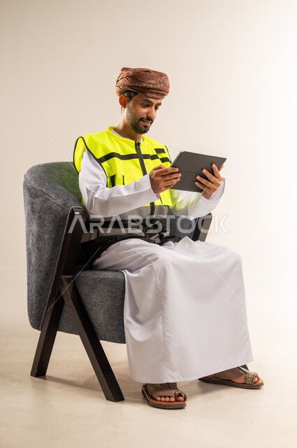 الإشراف والتدقيق على العمل، استخدام الاجهزة التقنية الحديثة، دمج التكنولوجيا الحديثة بالمجال الهندسي، بورتريه لرجل عربي خليجي عماني يرتدي سترة الحماية يجلس على كرسي مريح يحمل بيده التابلت، خلفية بيضاء