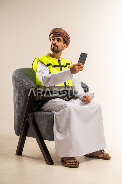 استخدام الاجهزة التقنية الحديثة في التدقيق والإشراف على العمل، دمج التكنولوجيا الحديثة بالمجال الهندسي، بورتريه لرجل عربي خليجي عماني يرتدي سترة الحماية يجلس على كرسي مريح يحمل بيده الهاتف المحمول ينظر الى مكان ما بايماءات الشرود، خلفية بيضاء