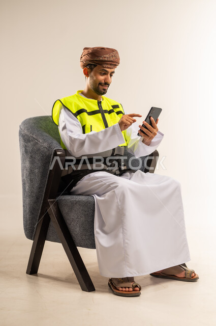 دمج التكنولوجيا الحديثة بالمجال الهندسي، استخدام الاجهزة التقنية الحديثة في التدقيق والإشراف على العمل، بورتريه لرجل عربي خليجي عماني يرتدي سترة الحماية يجلس على كرسي مريح يحمل بيده الهاتف المحمول، خلفية بيضاء