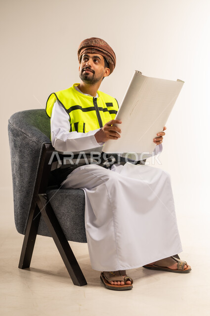 Engineering professions and jobs, auditing and supervising project plans, gestures of joy and happiness, portrait of an Arab Gulf Omani man wearing a work safety vest sitting on a comfortable chair holding an engineering plan in his hand looking at a place with an expression of distraction, white background