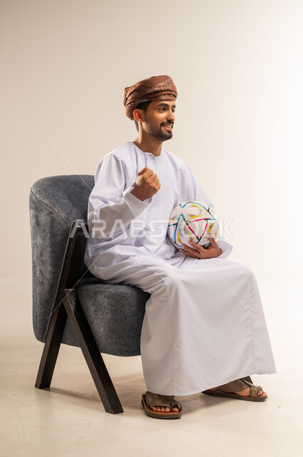 ايماءات السعادة بالانتصار والفوز، بورتريه من الجانب لرجل عربي خليجي عماني يرتدي الدشداشة والعمامة يجلس على كرسي مريح يحمل كرة قدم يقوم بمشاهدة المباريات يرفع يده بتعابير النصر، قضاء وقت ممتع في المنزل، الشغف في متابعه الفرق وتشجيع المنتخبات، خلفية بيضاء