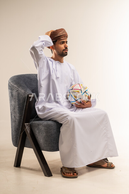 الشغف في متابعه الفرق وتشجيع المنتخبات، قضاء وقت ممتع في المنزل، بورتريه من الجانب لرجل عربي خليجي عماني يرتدي الدشداشة والعمامة يجلس على كرسي مريح يحمل كرة القدم يضع يده على رأسه بايماءات الحسرة والحزن للخسارة، خلفية بيضاء