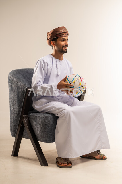 قضاء وقت ممتع في المنزل، ايماءات السرور والسعادة، بورتريه من الجانب لرجل عربي خليجي عماني يرتدي الدشداشة والعمامة يجلس على كرسي مريح يحمل كرة قدم يقوم بمشاهدة المباريات، الشغف في متابعه الفرق وتشجيع المنتخبات، خلفية بيضاء
