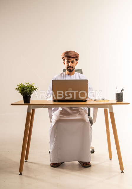العمل في المكاتب والشركات العمانية، دمج التقنيات الحديثة بالمجال الإداري والمكتبي، بورتريه لرجل عربي خليجي عماني يرتدي الدشداشه والعمامه يجلس خلف طاولة المكتب يقوم باستخدام الحاسوب المحمول في إنجاز المهام بإيماءات الاندماج والبشاشة، خلفية بيضاء