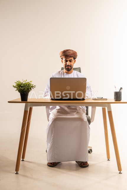 دمج التقنيات الحديثة بالمجال الإداري والمكتبي، العمل في المكاتب والشركات العمانية، بورتريه لرجل عربي خليجي عماني يرتدي الدشداشه والعمامه يجلس خلف طاولة المكتب يقوم باستخدام الحاسوب المحمول في إنجاز المهام بإيماءات الاندماج والبشاشة، خلفية بيضاء