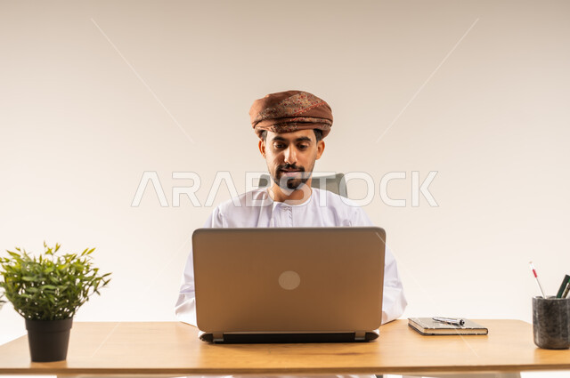 بيئة العمل في المكاتب والشركات العمانية، دمج التقنيات الحديثة بالمجال الإداري والمكتبي، بورتريه مقرب لرجل عربي خليجي عماني يرتدي الدشداشه والعمامه يجلس خلف طاولة المكتب يقوم باستخدام الحاسوب المحمول في إنجاز المهام بإيماءات الاندماج والبشاشة، خلفية بيضاء