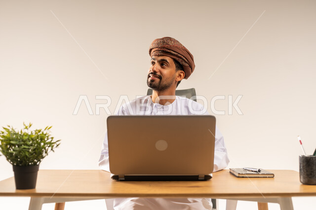 النظر إلى مكان ما بإيماءات الاندماج والبشاشة، العمل في المكاتب والشركات العمانية، دمج التقنيات الحديثة والتكنولوجية بالمجال الإداري والمكتبي، بورتريه مقرب لرجل عربي خليجي عماني يرتدي الدشداشه والعمامه يجلس خلف طاولة المكتب وأمامه الحاسوب المحمول، خلفية بيضاء
