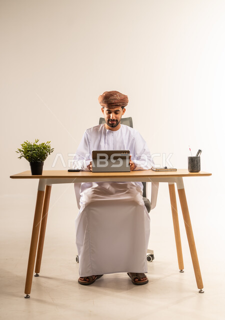 استخدام الجهاز اللوحي في الإشراف على سير الأعمال، مهن ووظائف في المكاتب والشركات العمانية، الاستفادة من التقنيات الحديثة في القطاع المكتبي والإداري، بورتريه لرجل عربي خليجي عماني يرتدي الدشداشه والعمامه يجلس خلف طاولة المكتب يدير العمل عن بعد، خلفية بيضاء