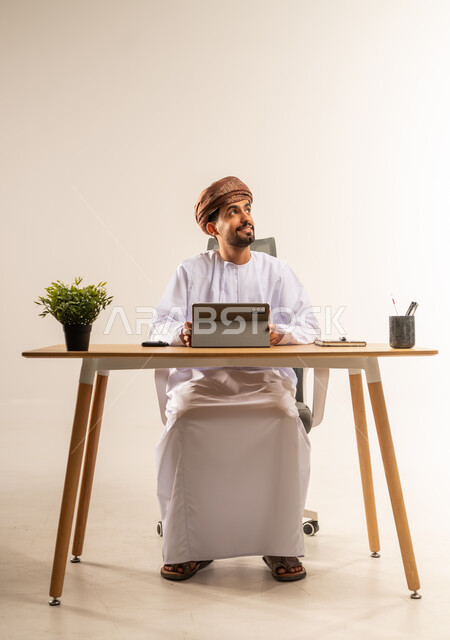 مهن ووظائف في المكاتب والشركات العمانية، الاستفادة من التقنيات الحديثة في القطاع المكتبي والإداري، الإشراف على سير الأعمال باستخدام الجهاز اللوحي، بورتريه لرجل عربي خليجي عماني يرتدي الدشداشه والعمامه يجلس خلف طاولة المكتب يدير العمل عن بعد، خلفية بيضاء