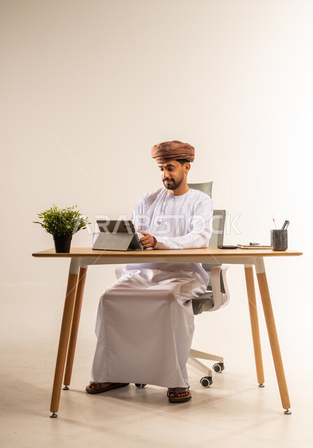 الاستفادة من التقنيات الحديثة في القطاع المكتبي والإداري، الإشراف على سير الأعمال باستخدام الجهاز اللوحي، بورتريه لرجل عربي خليجي عماني يرتدي الدشداشه والعمامه يجلس خلف طاولة المكتب يدير العمل عن بعد، مهن ووظائف في المكاتب والشركات العمانية، خلفية بيضاء