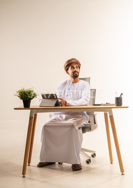 الإشراف على سير الأعمال باستخدام الجهاز اللوحي، الاستفادة من التقنيات الحديثة في القطاع المكتبي والإداري، بورتريه لرجل عربي خليجي عماني يرتدي الدشداشه والعمامه يجلس خلف طاولة المكتب يدير العمل عن بعد، مهن ووظائف في المكاتب والشركات العمانية، خلفية بيضاء