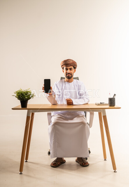 استخدام التكنولوجيا والتقنيات الحديثة المكاتب العمانية، النظر إلى الكاميرا بإيماءات البشاشة والسرور، إدارة سير الأعمال عبر الهاتف المحمول، بورتريه لرجل عربي خليجي عماني يرتدي الدشداشه والعمامه يجلس خلف طاولة المكتب ويعرض شاشة جوال سوداء فارغة، خلفية بيضاء