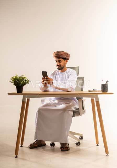 العمل في القطاع المكتبي والإداري، إدارة سير الأعمال عبر الهاتف المحمول، استخدام التكنولوجيا والتقنيات الحديثة المكاتب العمانية، بورتريه لرجل عربي خليجي عماني يرتدي الدشداشه والعمامه يجلس خلف طاولة المكتب يمسك الجوال بيده وبجانبه نبتة خضراء، خلفية بيضاء