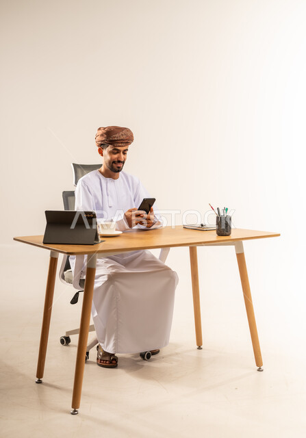 إدارة ومتابعة الأعمال عبر الهاتف المحمول، مفهوم الإشراف على العمل عن بعد، الاستفادة من التقنيات الحديثة في القطاع المكتبي، بورتريه من الجانب لرجل عربي خليجي عماني يرتدي الدشداشه والعمامه يجلس خلف طاولة المكتب يمسك الجوال بيده وبجانبه الجهاز اللوحي، خلفية بيضاء