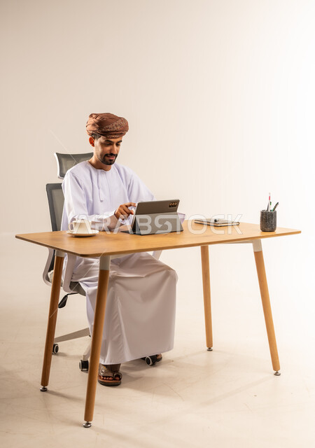 إدارة العمل عن بعد باستخدام الجهاز اللوحي، مهن ووظائف في المكاتب والشركات العمانية، الاستفادة من التقنيات الحديثة في القطاع المكتبي والإداري، بورتريه من الجانب لرجل عربي خليجي عماني يرتدي الدشداشه والعمامه يجلس خلف طاولة المكتب يعمل بجد وتركيز، خلفية بيضاء