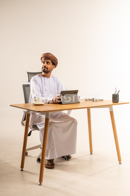 مفهوم إدارة العمل عن بعد، الإشراف على سير الأعمال عبر الجهاز اللوحي، الاستفادة من التقنيات الحديثة في القطاع المكتبي والإداري، بورتريه من الجانب لرجل عربي خليجي عماني يرتدي الدشداشه والعمامه يجلس خلف طاولة المكتب ينظر إلى مكان ما بإيماءات الاندماج، خلفية بيضاء