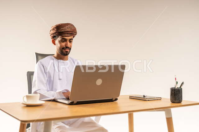 دمج التقنيات الحديثة والتكنولوجية بالمجال الإداري والمكتبي، تناول فنجان من القهوة الساخنة، العمل في المكاتب والشركات العمانية، بورتريه مقرب لرجل عربي خليجي عماني يرتدي الدشداشه والعمامه يجلس خلف طاولة المكتب ويعمل على جهاز الحاسوب المحمول بتركيز، خلفية بيضاء