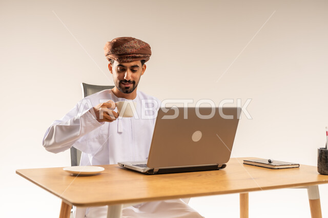 الاستمتاع بتناول فنجان من القهوة الساخنة، دمج التقنيات الحديثة والتكنولوجية بالمجال الإداري والمكتبي، العمل في المكاتب والشركات العمانية، بورتريه مقرب لرجل عربي خليجي عماني يرتدي الدشداشه والعمامه يجلس خلف طاولة المكتب وأمامه جهاز الحاسوب المحمول، خلفية بيضاء