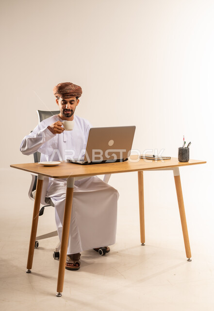 تناول فنجان من القهوة الساخنة، التركيز في إنجاز المهام، دمج التقنيات الحديثة بالمجال الإداري والمكتبي، العمل في المكاتب والشركات العمانية، بورتريه لرجل عربي خليجي عماني يرتدي الدشداشه والعمامه يجلس خلف طاولة المكتب ويعمل على جهاز الحاسوب المحمول، خلفية بيضاء
