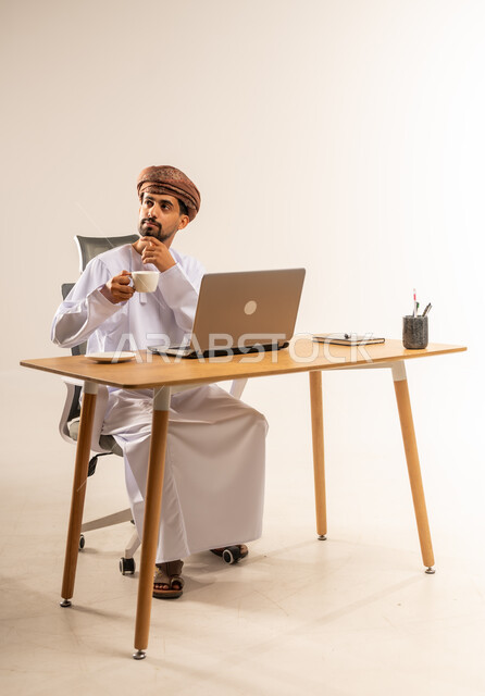 Deep thinking and contemplation while drinking a cup of hot coffee, integrating modern technologies into the administrative and office field, working in Omani offices and companies, portrait of an Arab Gulf Omani man wearing a dishdasha and turban sitting behind an office table with a laptop in front of him, white background