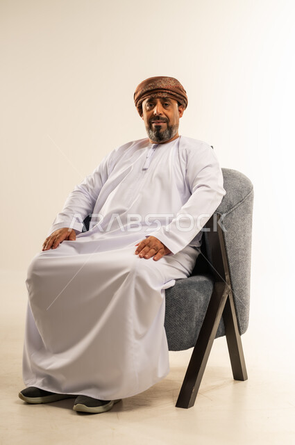 الاهتمام بالمظهر الخارجي، مفهوم الرجولة والوقار، ايماءت الثقة بالنفس، بورتريه لرجل عربي خليجي عماني يرتدي الدشداشة والعمامة يجلس على أريكة مريحة ينظر الى مكان ما بتعابيرالسرور، خلفية بيضاء